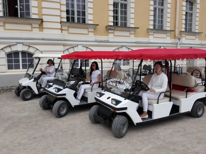 Trīs baltie elektromobiļi ar uzrakstu „Ekskursija pa parku” gaida pie Rundāles pils dzeltenās fasādes.