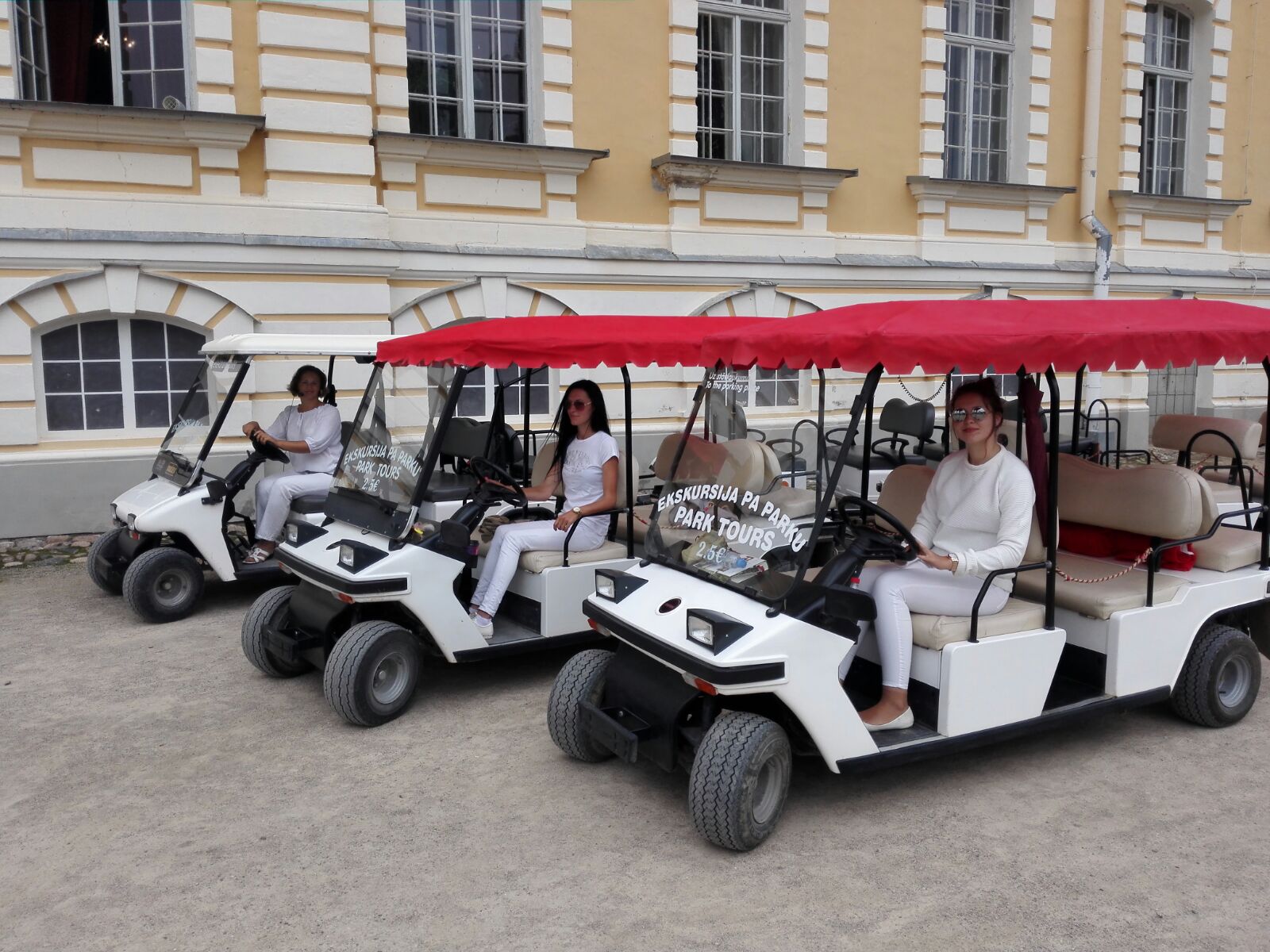 Trīs baltie elektromobiļi ar uzrakstu „Ekskursija pa parku” gaida pie Rundāles pils dzeltenās fasādes.