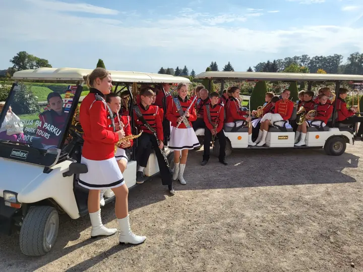 Jauniešu pūtēju orķestris sarkanos tērpos pie elektromobiļiem Rundāles pils parkā.