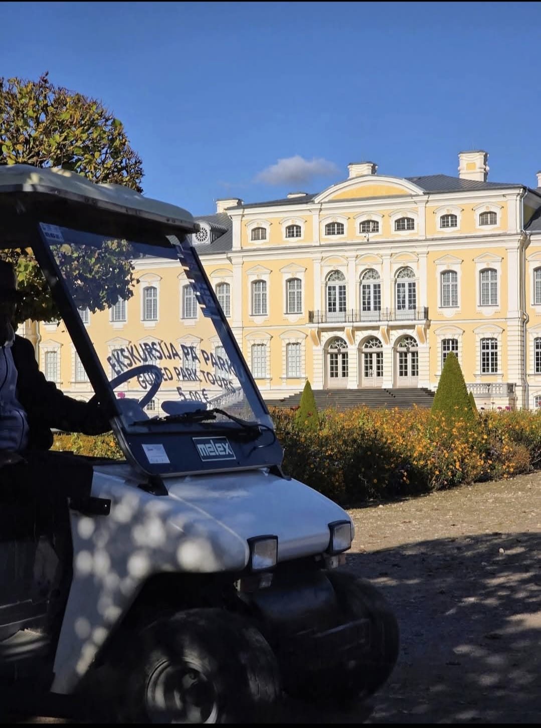 Elektromobilis „Ekskursija pa parku” pie Rundāles pils dzeltenās fasādes rudens saulē.